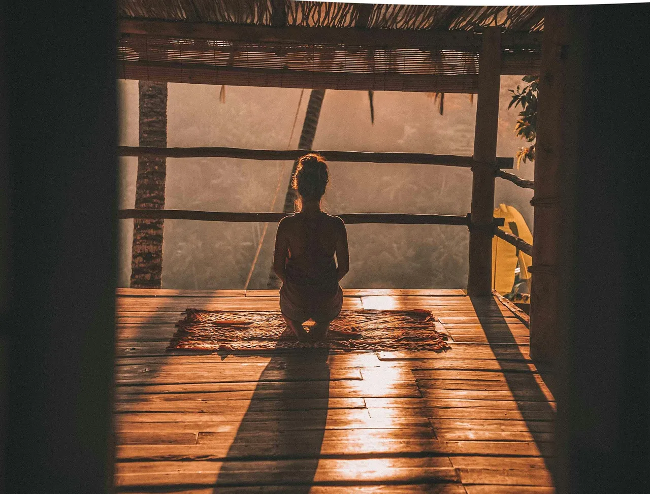A person kneeling in silhouette on a mat on a wooden deck, facing out into misty trees at sunrise