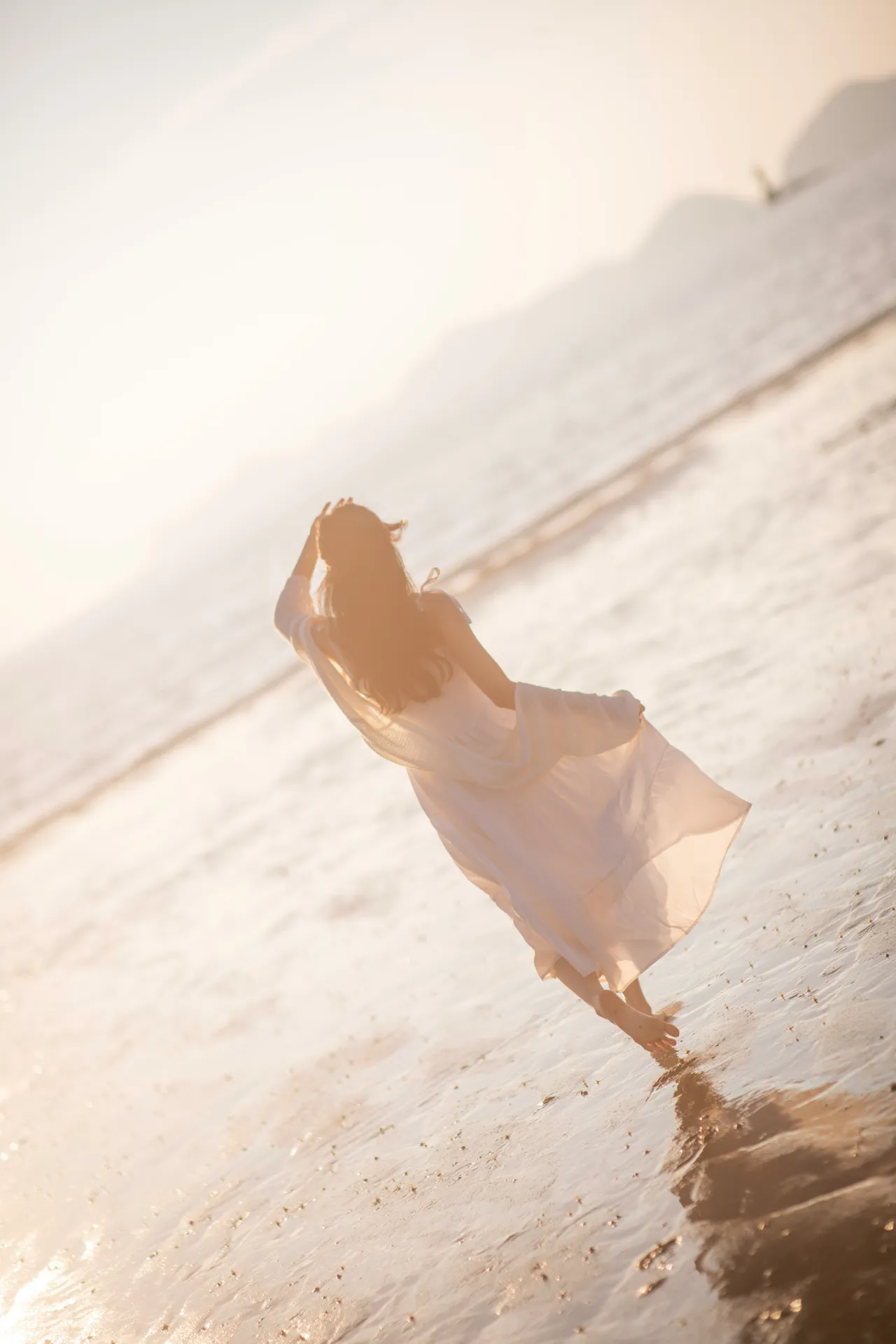 A person in a pale flowing dress walking barefoot along a sunlit shoreline